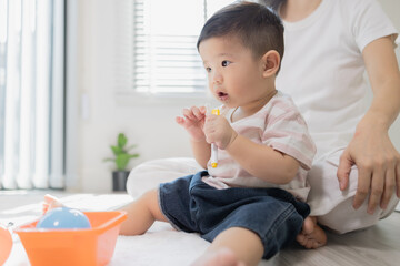 Fototapeta premium Warm sunlight bathes a cozy playroom scene. Mother gently holds her infant, eyes full of affection. Soft toys and bright containers dot the floor nearby. Natural light filters through window blinds 