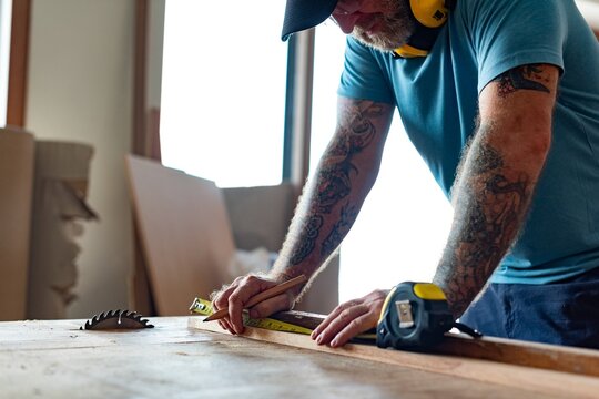 Man measuring wood in a workshop, wearing a cap and ear protection.