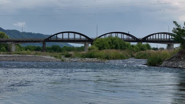 River flows beneath three arched bridges. Greens and stones line riverbanks. Clouds drift above distant hills. No people are present. Perfect for tranquil landscape projects