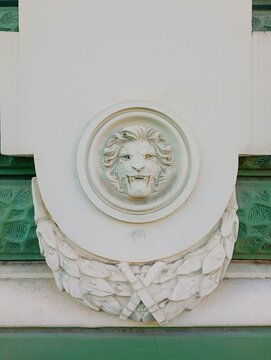 Close-up photograph of a classical architectural mascaron featuring a carved lion head medallion with a laurel wreath garland on a building facade. High-quality historical exterior image ideal for arc