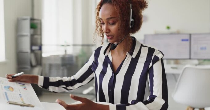 Woman operator agent with headset handles call support. She works in a bright office, reviewing data and assisting clients by phone with sales, service and tech issues. Clear care concept.
