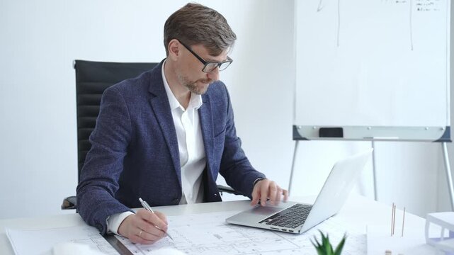 Architect or engineer working on construction blueprints and plans at an office desk, using a laptop for design and planning, embodying professionalism and project management