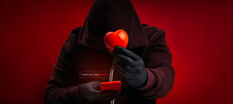 Hooded scammer holding warning heart ball, using smartphone and fake emotional message for online romance scam. Catfishing, emotional manipulation, dating fraud, cybercrime, digital deception.