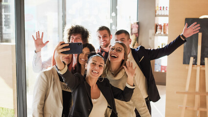 A group of friends stands together in a modern cafe during the afternoon. They are smiling and posing for a selfie. Some wave at the camera © Nikola Spasenoski