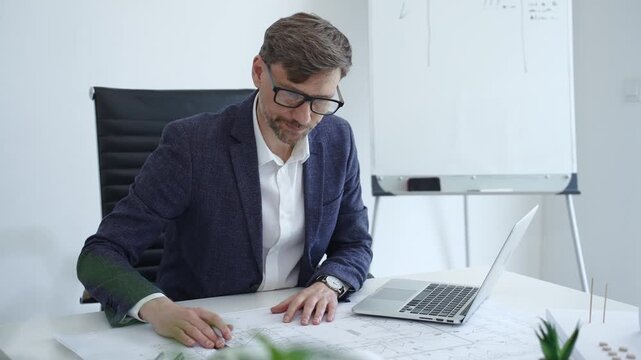 Architect or engineer working on construction blueprints and plans at an office desk, using a laptop for design and planning, embodying professionalism and project management