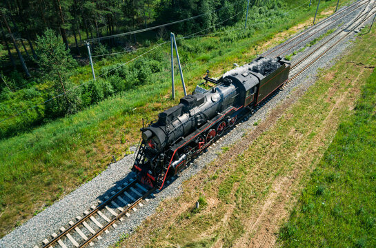Aerial view captures an old black steam locomotive moving along parallel train tracks next to a dense green forest