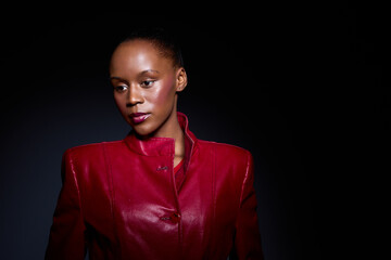 Obraz premium Elegant dark skinned African woman in red jacket poses against black background. Studio portrait with dramatic lighting highlights her confident expression and fashion style.