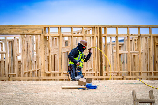 Construction worker holding air hose and pneumatic nail gun while standing on OSB roof of wood-framed building