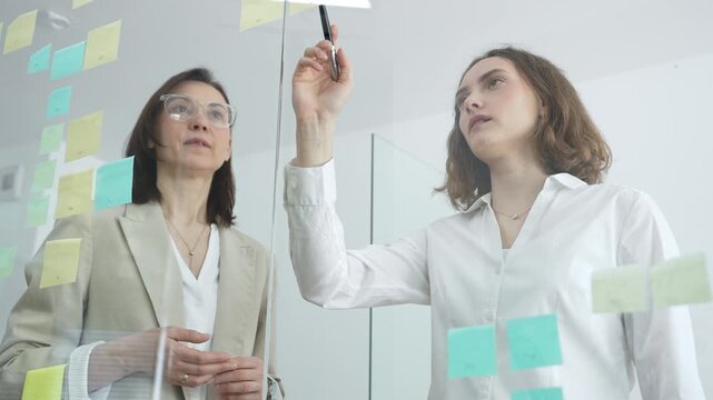 Diverse businesswomen collaborating and brainstorming innovative ideas while sticking and arranging colorful post it notes on a clear glass wall in a modern office meeting or workshop