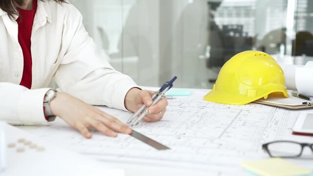 Female architect or engineer working on blueprints using a compass and ruler. Designing building plans. With a yellow hard hat and tablet on the desk