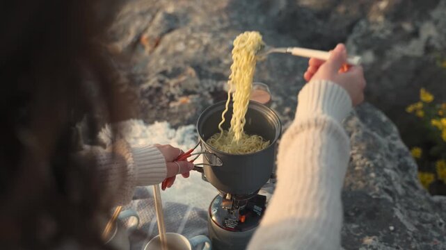 A woman cooks noodles on a portable stove outdoors on rocks