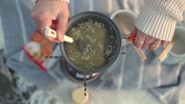 Instant Noodle Comfort: Hands stirring a pot of steaming instant noodles, a simple and comforting meal prepared outdoors.