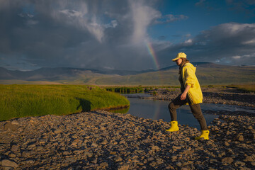 Fototapeta premium Side view of young woman in yellow rain boots hiking near creek with rainbow in sky. Female traveler exploring remote mountain valley landscape after storm during golden hour.