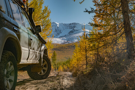 Side view of offroad SUV car parked on dirt road in autumn forest. Expedition 4x4 vehicle traveling through remote scenic mountain wilderness with snowy peak under blue sky.