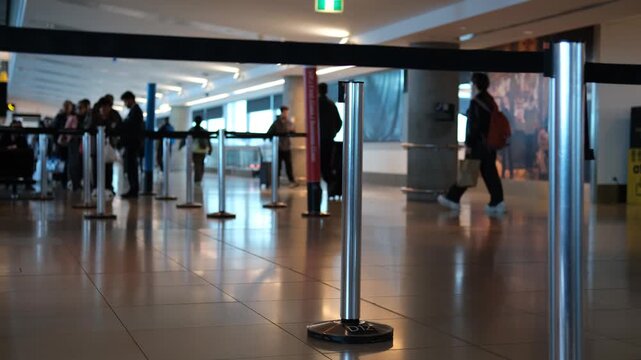 Close-up on a chrome retractable belt queue barrier stanchion in foreground of a busy airport terminal, with blurred passengers and travellers moving through concourse in a busy international airport