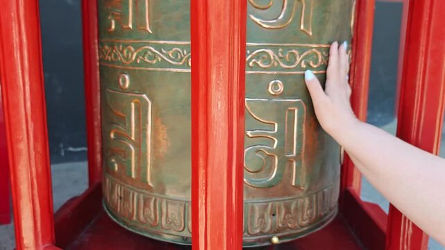 A womans hand with blue nails turns a large bronze prayer wheel set in a red wooden frame adorned with intricate carvings and bright flowers, against a backdrop of serene nature