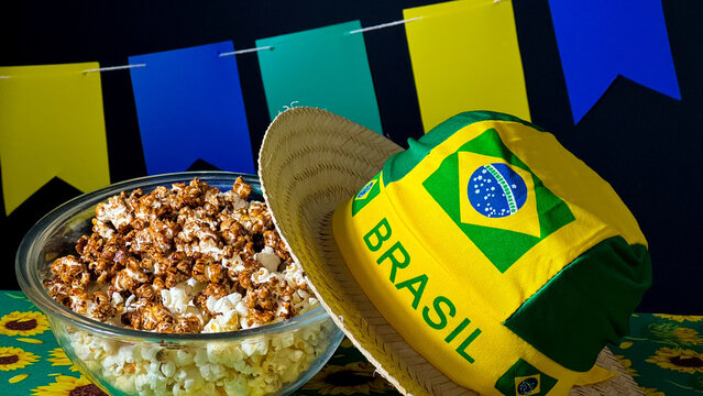 A bowl of popcorn, a straw hat, a scarf with the Brazilian flag symbol and the word "Brasil" (in Portuguese), and colorful flags in the background. Traditional Brazilian June festival. Arrai&aacute;.