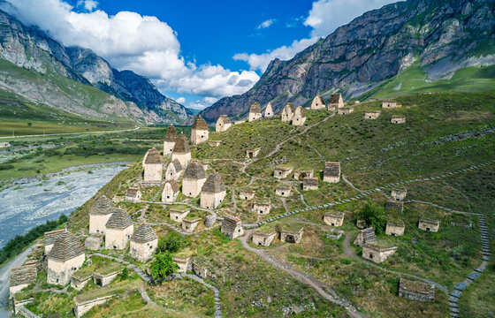 Aerial view captures Dargavs City of the Dead sit on a hill near a river with mountains in the background during a sunny day in North Ossetia-Alania