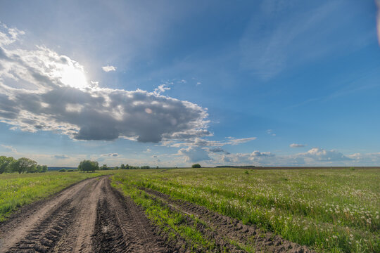 Rural dirt road through green summer field under dramatic cloudy blue sky