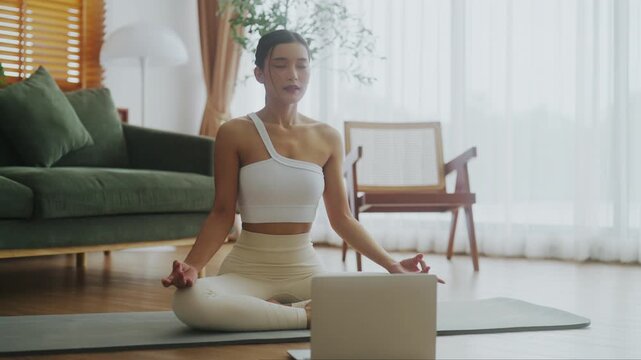 Happy young Asian woman practicing yoga and meditation at home sitting on floor in living room in lotus position and relaxing. Mindful meditation, Wellbeing, Breathing exercise