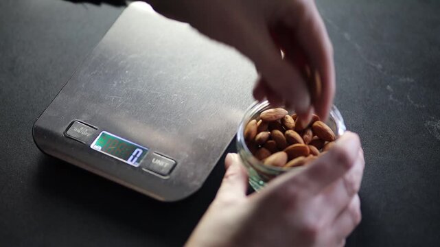 Close-up video showing hands counting almonds and placing them one by one onto electronic kitchen scales in a home kitchen setting. The scene highlights careful portion control.