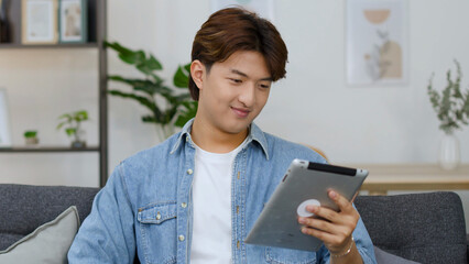 Young Asian Man Smiling While Using a Digital Tablet on a Sofa © blueringmedia