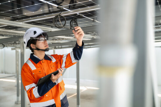 Young engineer in orange uniform and white helmet inspects solar panels, checking cable connection point under solar panels while holding a tablet check power consumption received via tablet.