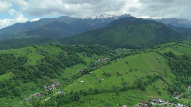 Epic sweeping panoramic view of Simon village and fresh green hills under a moody sky with snowy Bucegi mountains in background, high altitude drone pan