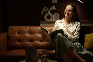 Fototapeta premium Smiling young woman reading fashion magazine on comfortable leather sofa at home, enjoying evening relaxation and leisure