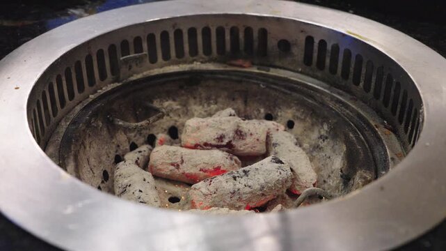 Close up of glowing charcoal embers in a round metal tabletop grill at a restaurant. Metal tongs adjusting burning coals for Japanese yakiniku or Korean barbecue cooking. Intense heat. High angle.