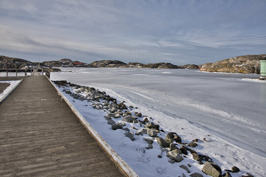 Symmetrical view of a wooden pier with rope railings leading across the ice in Skrhamn. A cold winter day in the Swedish archipelago with dramatic light and frozen wate