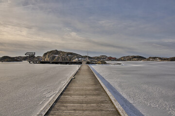 Obraz premium Symmetrical view of a wooden pier with rope railings leading across the ice in Skrhamn. A cold winter day in the Swedish archipelago with dramatic light and frozen wate