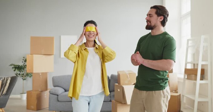 Couple moving, woman shows surprise and reaction. Man presents a bright apartment with moving boxes, marking the reveal of a new home and pure joy of settling in. Concept of new home joy