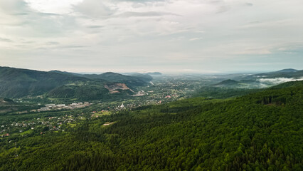 Fototapeta premium Expansive view from forested hills toward a sprawling valley town. Rolling ridges frame neighborhoods roads and fields while wisps of fog linger in sheltered folds beneath an overcast summer sky.