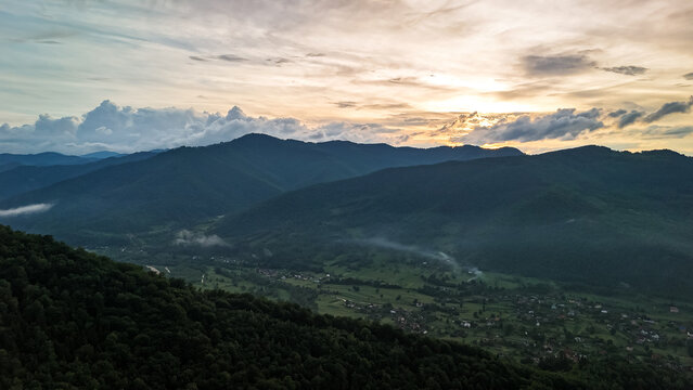 Soft sunset light spreads over a mountain valley where specks of smoke and fog linger above fields. Dark slopes frame the scene as fading colors ripple across a sky of layered clouds.