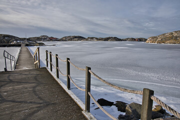 Naklejka premium Symmetrical view of a wooden pier with rope railings leading across the ice in Skrhamn. A cold winter day in the Swedish archipelago with dramatic light and frozen wate