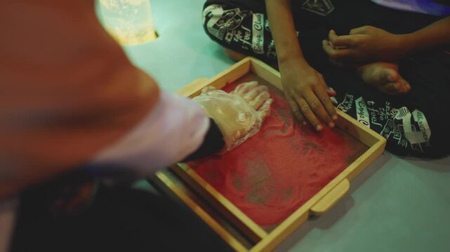 Hands of young girls collaborating and drawing patterns inside a wooden sandbox table at a special school. Static close-up.