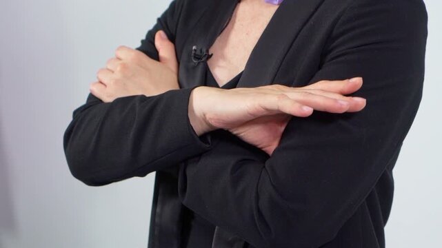 Close-up of a person&rsquo;s upper torso with arms crossed, wearing a black blazer and dark top. A lavalier microphone is clipped near the neckline. Both hands are resting on the opposite arms