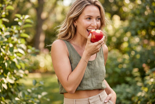 Young woman taking a bite of a fresh red apple in a green outdoor garden setting