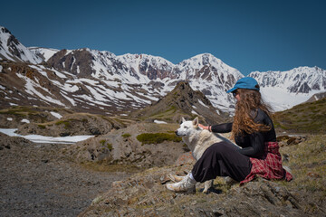 Side view of young woman sitting and petting her white dog on mountain hill. Female traveler with husky exploring remote scenic wilderness landscape under beautiful blue sky. © ANDREY