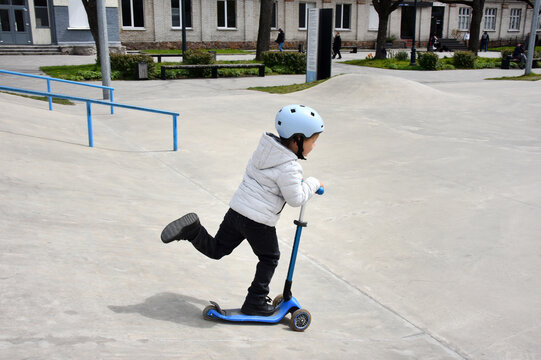 Young boy riding a blue scooter at a skatepark wearing a helmet and