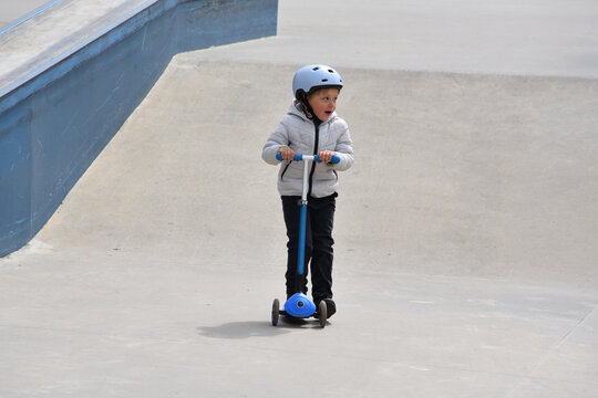 Young boy riding a blue scooter at a skatepark wearing a helmet and