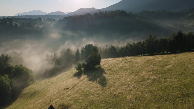 Dynamic timelapse of rapidly moving morning mist in a rural mountain valley with striking sunbeams through trees, scenic golden hour in the Bran hills, Carpathians, static aerial view