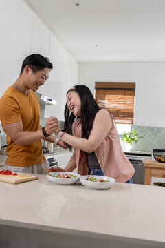 Asian couple laughing and preparing salad at kitchen island with white bowls and tomatoes