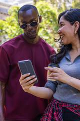 Fototapeta premium Diverse couple leaning showing phone holding coffee wearing shades and plaid tied at waist in plaza