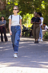 Obraz premium Woman walking toward camera on park walkway wearing sunglasses, backpack, cargo jeans and sneakers