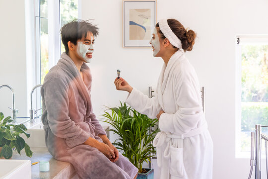 Diverse couple wearing plush bathrobes applying facial masks at bathroom countertop with brush, jar