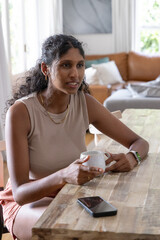Adult Indian woman in sleeveless beige top sitting at table at home holding white mug, smartphone © wavebreak3
