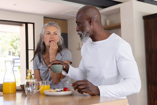 Diverse couple sitting at kitchen island tasting pancakes with berries near carafe of orange juice