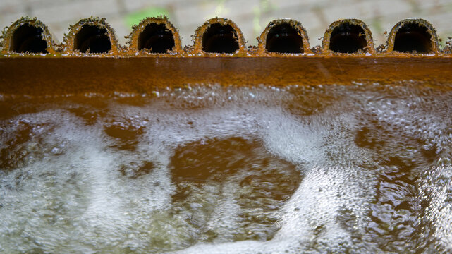 Muddy water bubbling inside a rusty metal industrial filtration or settling tank with a row of overflow pipes, close-up.